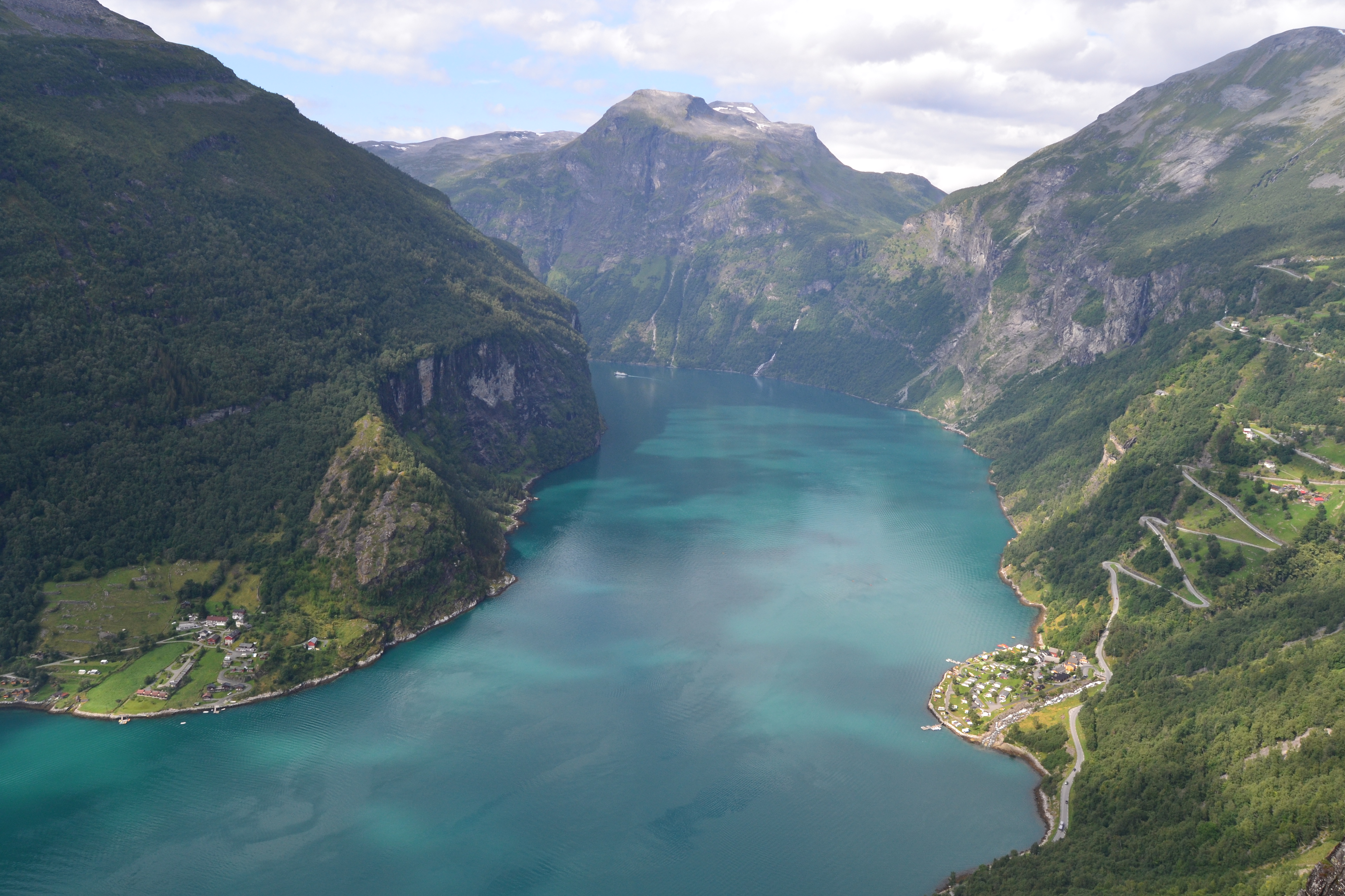 Panoramic view from Løsta viewpoint 500 meters above Geirangerfjord showing turquoise water, Geiranger village, and Eagle Road hairpins