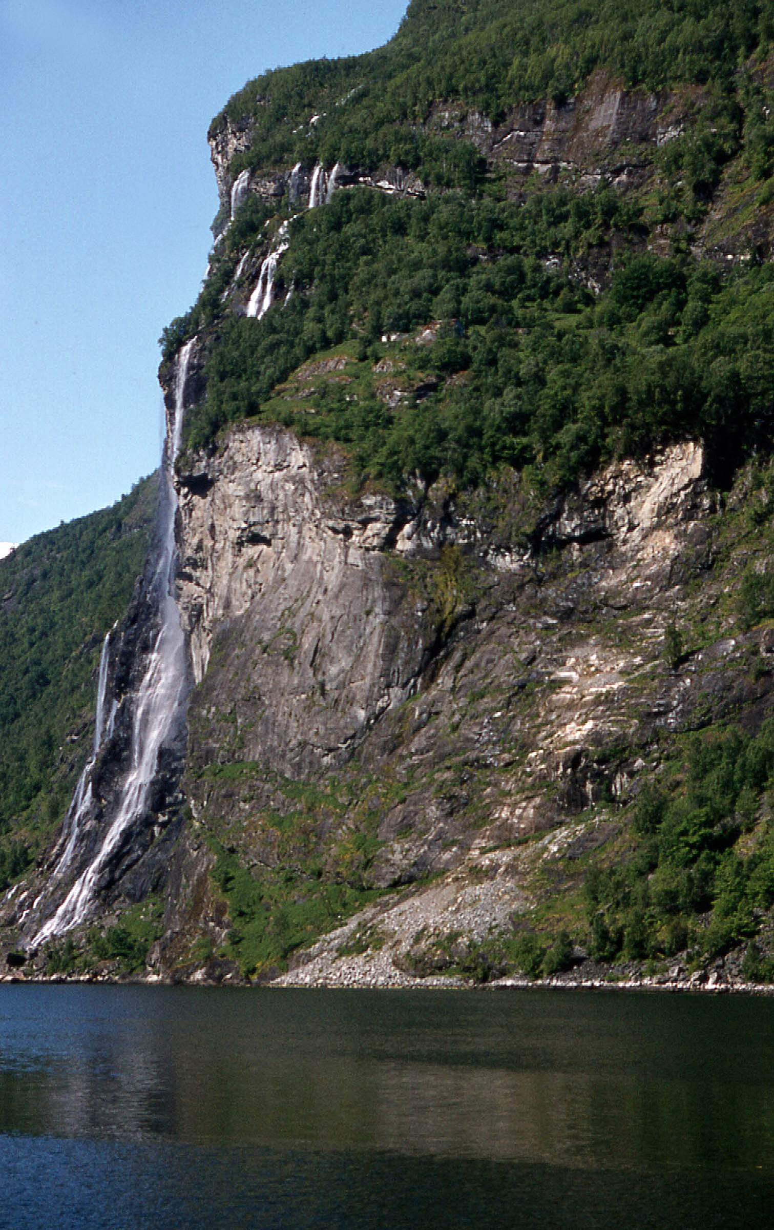 The Seven Sisters waterfall cascading 250 meters down the steep cliff face of Geirangerfjord, viewed from water level with the rock wall towering above