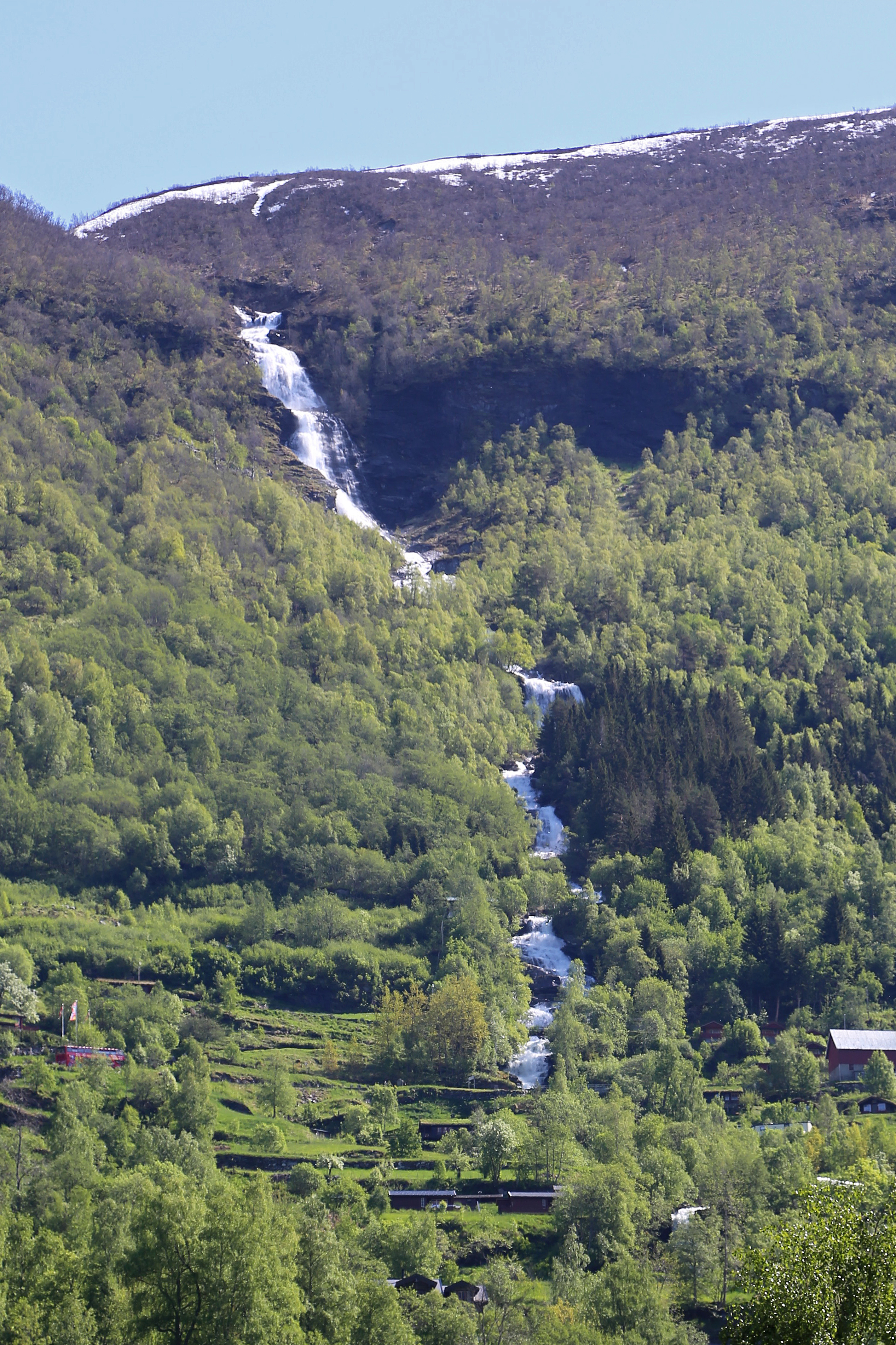 Storseterfossen waterfall cascading down a forested mountainside above Geiranger village, river flowing through green birch forest with snow on the plateau above