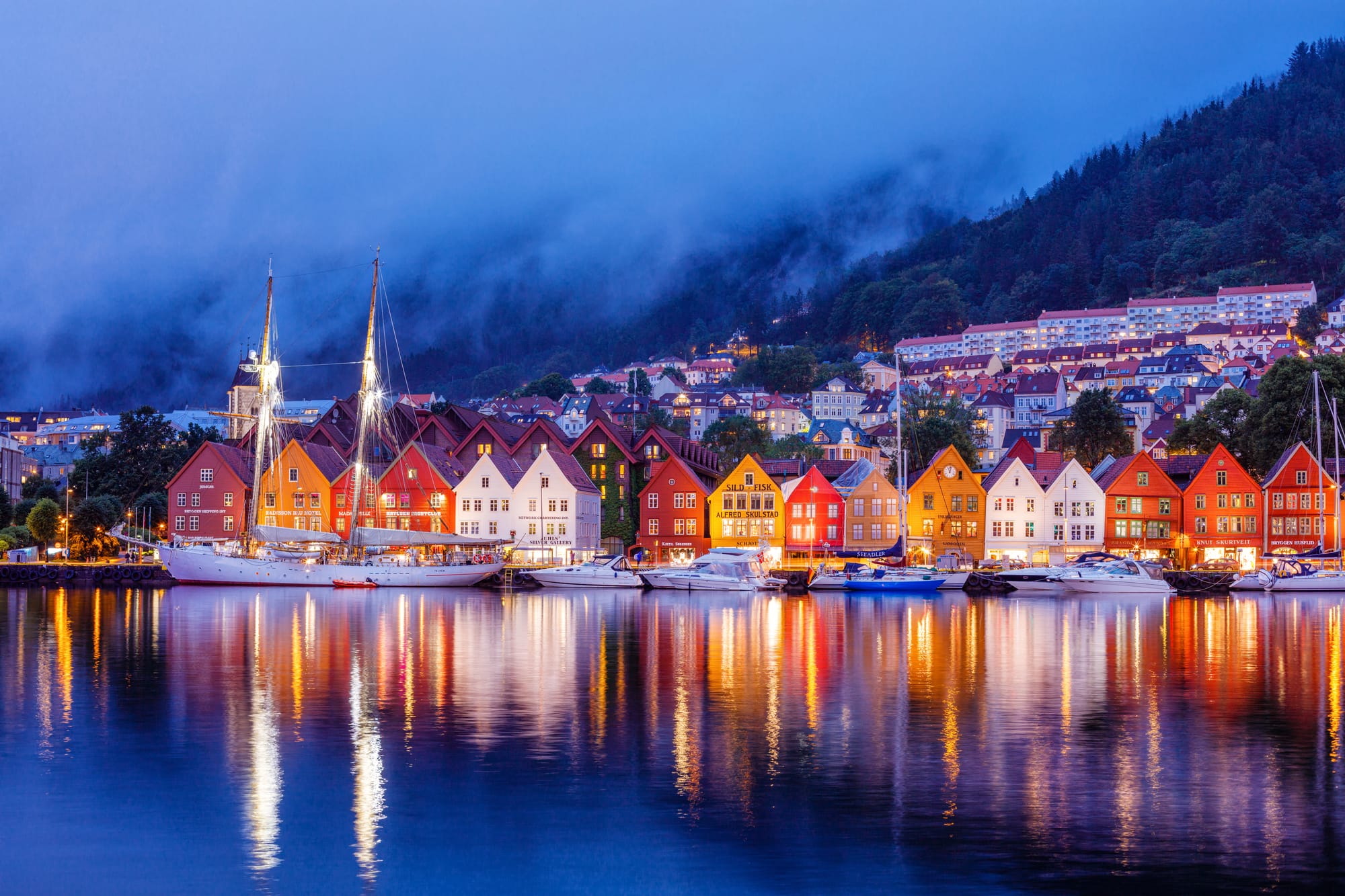 Bryggen wharf houses on the Bergen harbour front lit at dusk, low cloud on the wooded hillside behind and boats moored along the quay